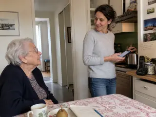 Senior woman in a Nantes kitchen chats naturally with her adult daughter, smartphone in hand, in gentle daylight, with handwritten notes and a cordless phone nearby.