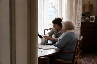 Senior woman and her daughter setting up a tablet together at a sunlit kitchen table, notes and tea nearby, captured from the doorway with natural, candid interactions.