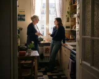 Senior woman in a Nantes kitchen prepares tea while conversing with a younger neighbor, soft natural light streaming through window, calm and relaxed atmosphere.