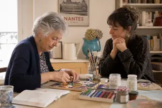 Senior woman and adult daughter at home in Nantes, collaborating on a mosaic art project at the dining table in natural daylight, surrounded by art materials.