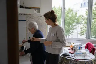 Elderly woman and volunteer cooking together in a sunlit Nantes kitchen, interacting naturally