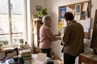 Senior woman and middle-aged man side-by-side in a sunlit Nantes apartment, thoughtfully looking at a community noticeboard with local event flyers, everyday home details visible in a gentle, candid scene.