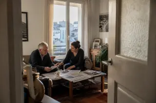 Senior man and adult daughter in a sunlit Nantes living room sharing an attentive conversation at a cluttered coffee table, viewed candidly from a doorway