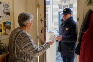 Senior woman and local postal worker exchange a note in a softly lit apartment entryway, warmth and trust visible in a candid, natural moment.