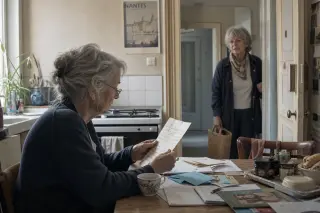 Elderly woman reading a handwritten letter at a kitchen table, with a neighbour pausing in the doorway behind her, in softly lit Nantes apartment