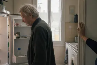 Senior man in casual clothes looks into an open fridge in a softly lit kitchen, while a woman’s hand on the doorframe in the background offers subtle support, natural midday light coming from a window