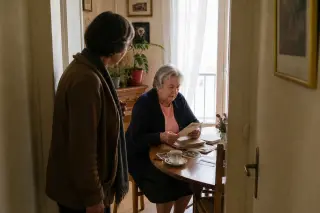 Senior woman sorts through letters at her kitchen table while a neighbor quietly checks in from the doorway, natural light creating a soft, intimate atmosphere.