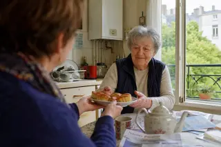 Senior woman at kitchen table receiving pastries from a neighbor in a warm, softly lit Nantes apartment