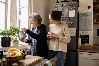 Senior woman and her adult daughter quietly watering plants together in a natural, sunlit Nantes kitchen, showing a calm, authentic moment.