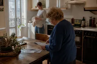 Senior woman sorting mail at a kitchen table with a caring adult nearby in a sunlit Nantes apartment, natural and unposed atmosphere.
