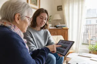 Senior woman in a Nantes living room teaching a young volunteer to organize photos on a tablet, natural daylight and relaxed ambiance, both focused on the screen.