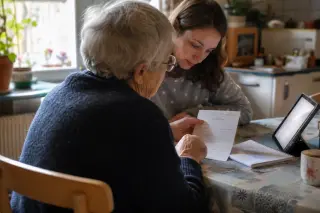 Senior woman and younger helper study a handwritten guide together at a kitchen table, tablet nearby, natural daylight creating a warm, lived-in atmosphere.