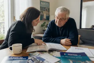 Senior man and adult daughter sitting at a table in a Nantes apartment, discussing municipal service brochures in soft daylight, focused and absorbed.