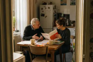 Senior man and middle-aged woman, in a cozy Nantes kitchen, discuss over training documents and tea, bathed in soft afternoon light.