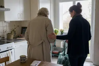Senior and adult daughter in a softly lit kitchen, preparing together for a local outing, sharing a subtle supportive moment.