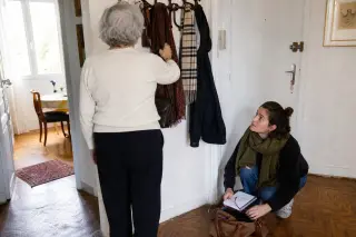 Senior woman selecting a scarf by the door in a sunlit apartment, while a younger helper organizes a bag nearby, both engaged in a calm, everyday scene.