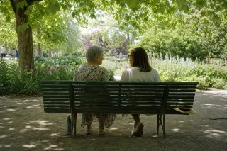 Two women, one senior and one middle-aged, sitting on a green bench under trees in a Nantes park, sunlight filtering through leaves, chatting quietly.