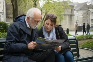 Senior man and middle-aged woman looking at a city brochure together on a park bench in Nantes, soft spring light, relaxed and natural atmosphere.
