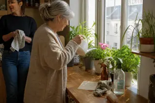 Senior woman watering plants on a kitchen windowsill, soft sunlight, domestic scene, adult daughter in background tidying up.