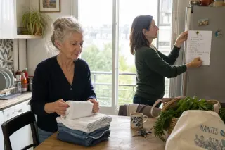 Senior woman folding towels in a Nantes apartment kitchen as her daughter checks a household task list, natural lighting and calm atmosphere
