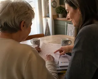 Senior woman and adult daughter reviewing notes together at a sunlit kitchen table, warm and natural atmosphere.
