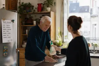 Senior man cleaning a kitchen counter with his adult daughter nearby, natural light entering through a window, everyday home atmosphere in Nantes.