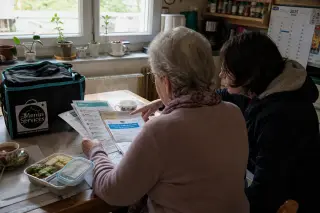 Senior and family member in Nantes kitchen calmly sorting delivery meal documents with warm food bag on table, natural light creating a cozy, authentic moment.