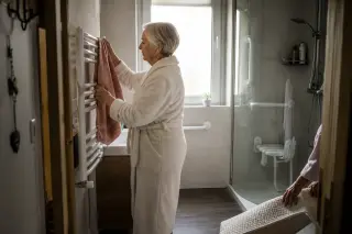 Senior woman in bathrobe arranging towel in a softly lit bathroom as her daughter places a non-slip mat by the shower, showing quiet morning routine and safety features in a real Nantes home.