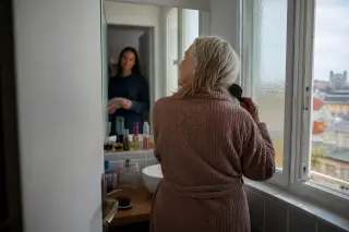 Senior woman brushing hair in bathroom, daughter waits in hallway with towel, cozy natural light, intimate and respectful atmosphere