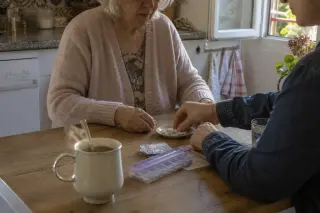 Senior woman and younger adult at a wooden kitchen table, hands interacting with medication, morning light, homely Nantes kitchen