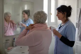 Senior woman and caring aide in a real Nantes bathroom, preparing for a gentle morning wash together, reflected in a misted mirror, natural light, unstaged atmosphere.