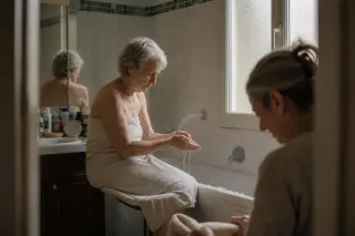 Elderly woman sitting at the edge of a bathtub in a softly lit bathroom, washing her hands while her daughter quietly prepares a towel nearby.